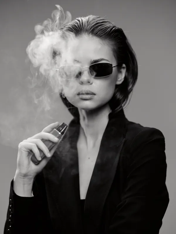 Black and white portrait of a stylish woman vaping in sunglasses with smoke around her face.