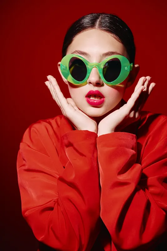 Stylish young woman wearing oversized green sunglasses and rich red lipstick poses with surprise on vivid red background. Fashion and beauty concept with vibrant colors and bold expression.