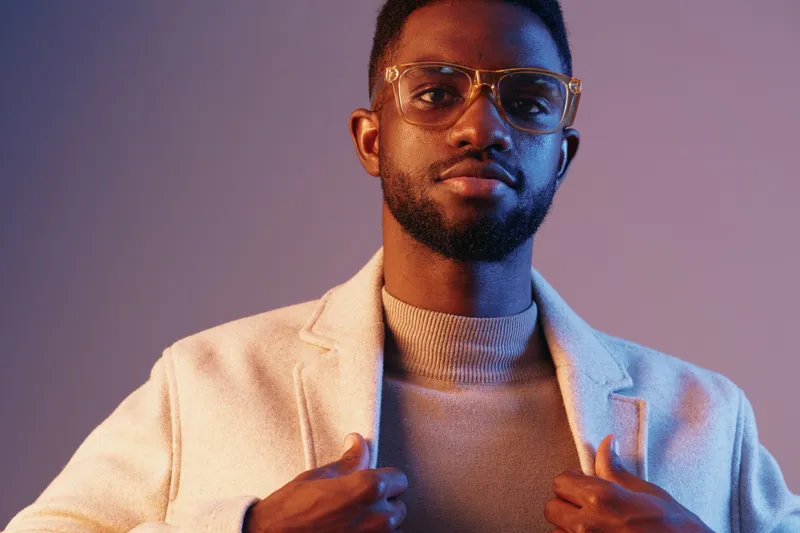 Stylish young man wearing glasses and a light coat, showcasing confidence against a purple gradient background, ideal for fashion promotions and modern lifestyle imagery