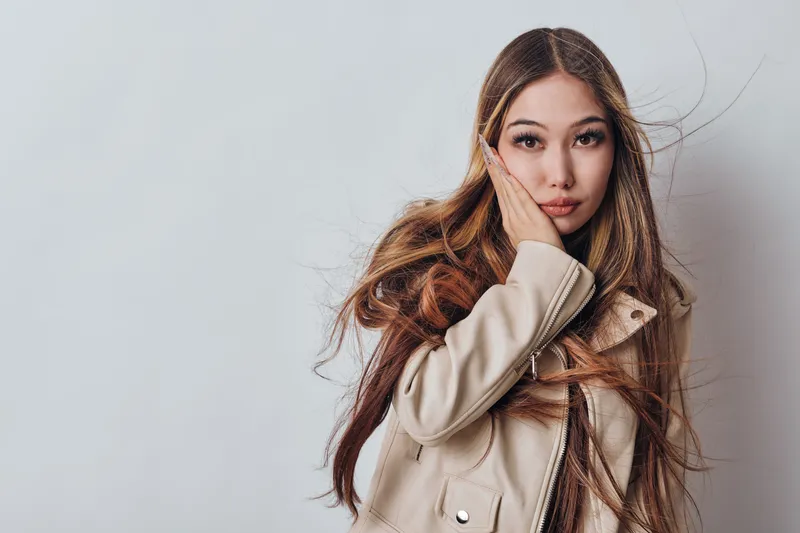 Young woman with long hair in coat poses with hand on face against plain background. Fashion model looking at camera with serious expression in studio portrait.