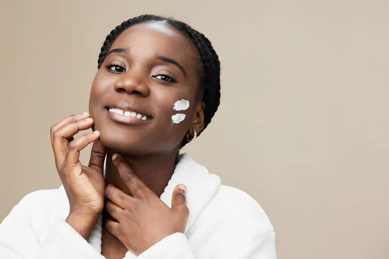 Smiling Black woman applying skincare on her face, wearing a white robe, with a neutral beige background representing healthy skincare routine