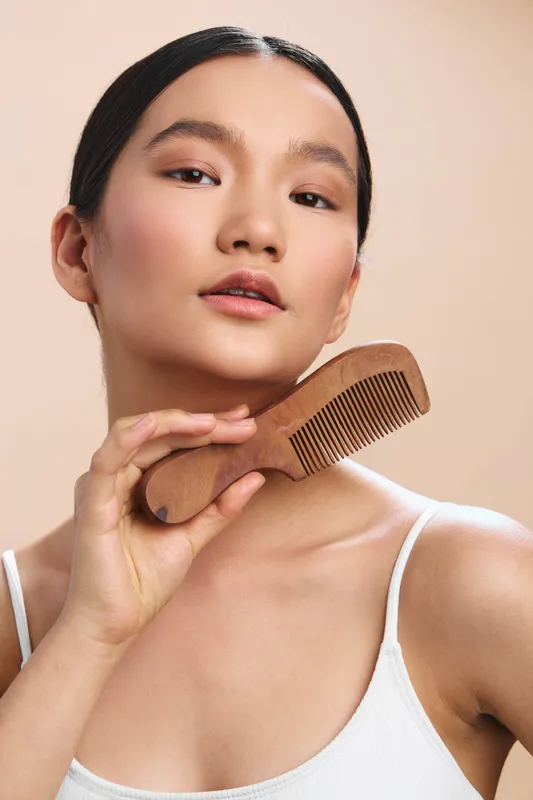 asian woman is photographed in a soft studio setting, gracefully holding a wooden comb as she styles her hair. the calm expression and gentle light create a natural, confident beauty mood.