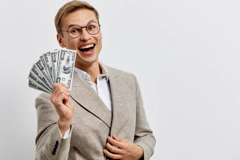 Confident smiling woman with short hair and glasses holding a fan of dollar bills in her hand, wearing a beige blazer over white shirt, business success concept, studio portrait with plain white backg