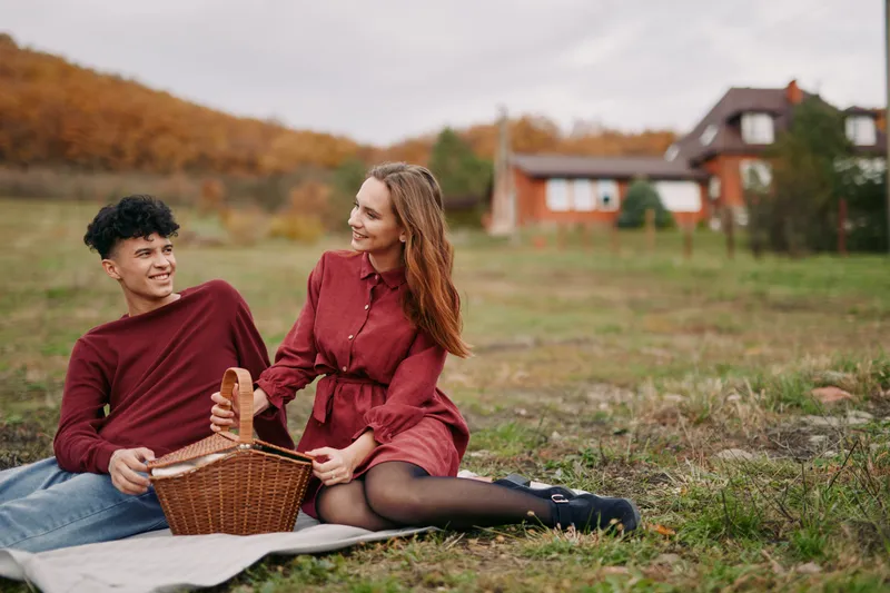 A warm authentic picnic scene of a smiling couple sitting on a blanket with a wicker basket by a cottage, genuine interaction and natural expressions capturing a trustworthy, real relationship moment.