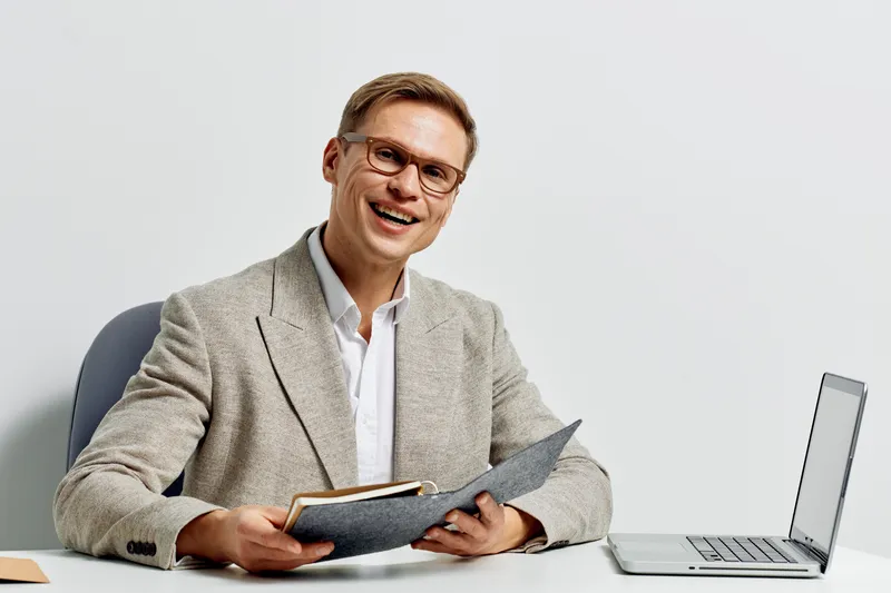 Smiling confident man in business attire sitting at desk with laptop and documents, working in modern office environment, professional, cheerful expression, light background, concept of work and succe