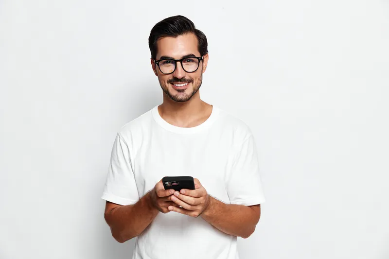 Smiling young man wearing glasses holding smartphone looking at camera isolated on plain solid light background. Modern lifestyle concept.