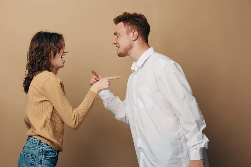 Young man and woman argue facing each other with intense expressions on beige background. Concept of conflict, disagreement, communication, and relationship tension between couple.