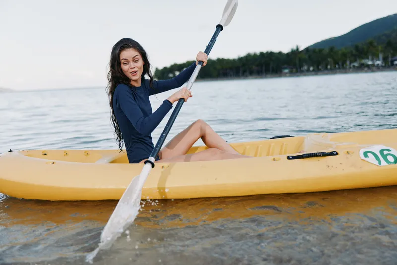 A young woman paddles a bright yellow kayak on calm coastal water, gripping a double blade paddle as she glides gracefully along the sandy shore.