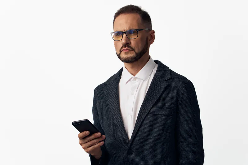 A confident business professional man stands against an isolated white background, wearing a textured blazer and white shirt, holding a smartphone, exuding focus and resilience.