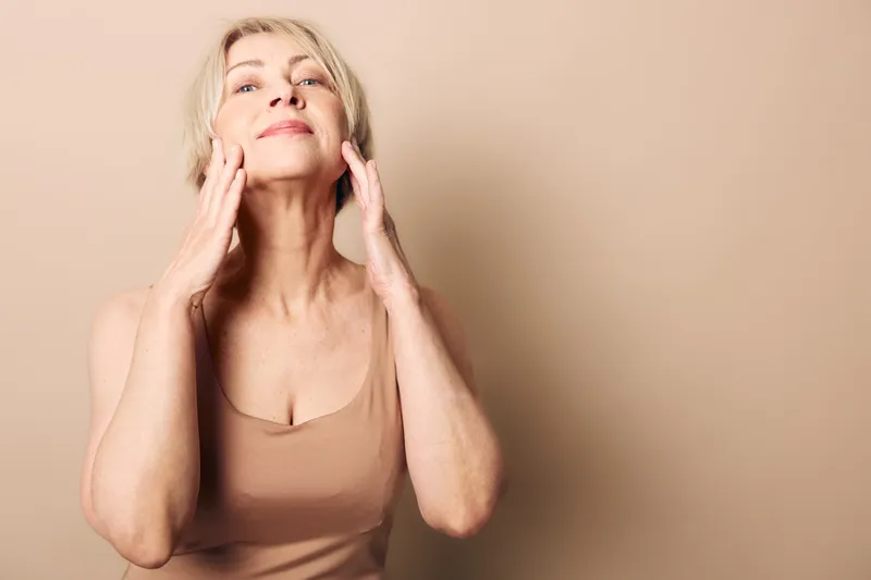 Mature woman with light hair applies skincare cream on face, wearing beige top. Soft neutral background highlights beauty, self-care, and natural wellness concept for aging skin.