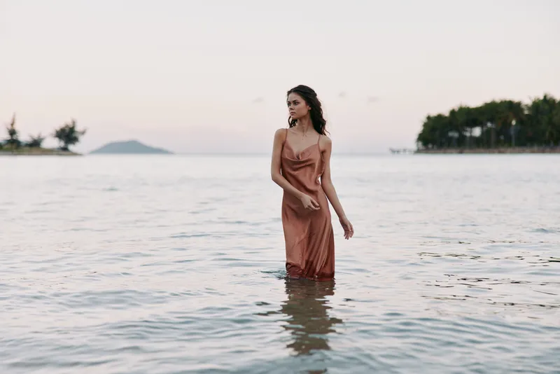 A woman in a silky rust-colored dress stands knee-deep in the calm sea at sunset, creating a serene, graceful portrait with gentle waves and reflective light.
