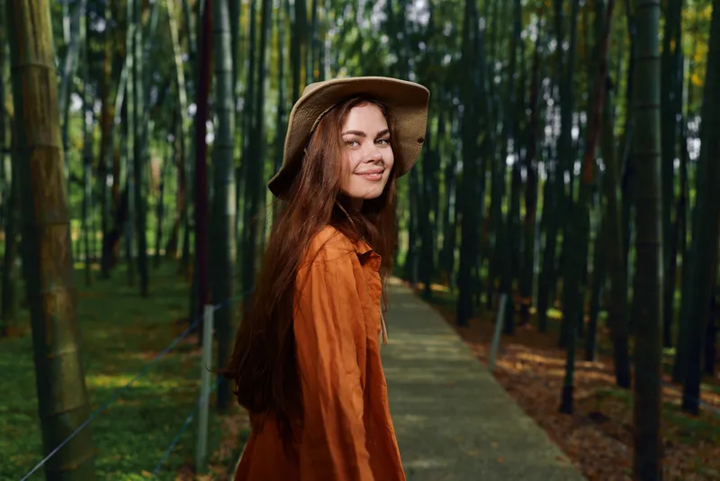 Woman smile hat bamboo forest path portrait walking in nature, young female with long hair in orange coat looking back on wooden walkway surrounded by green trees and sunlight.