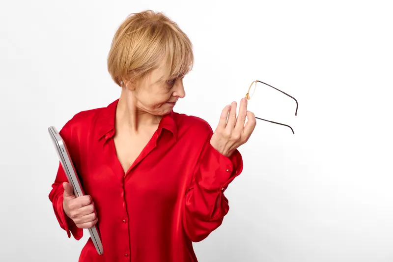 Elderly woman in red shirt holding book and glasses, making a confused gesture while looking away, senior mature expression, white studio background, close portrait.