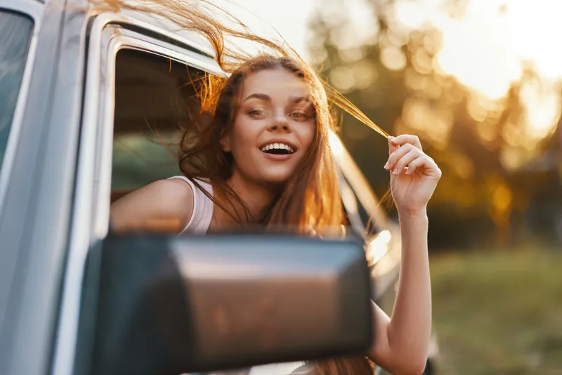 Young woman enjoying a road trip with wind in her hair, smiling from a car window during sunset, capturing a sense of freedom and adventure.