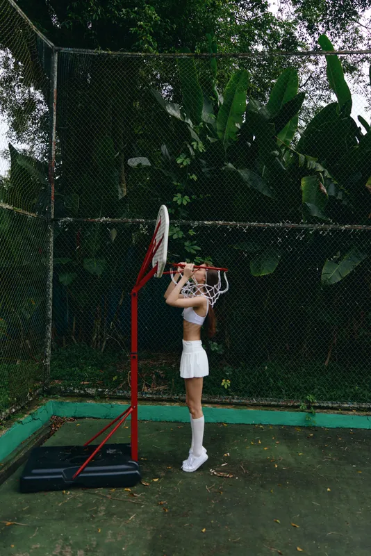 Young female basketball player in a white sports outfit stands by the hoop in a lush outdoor setting, showcasing a moment of concentration and athleticism