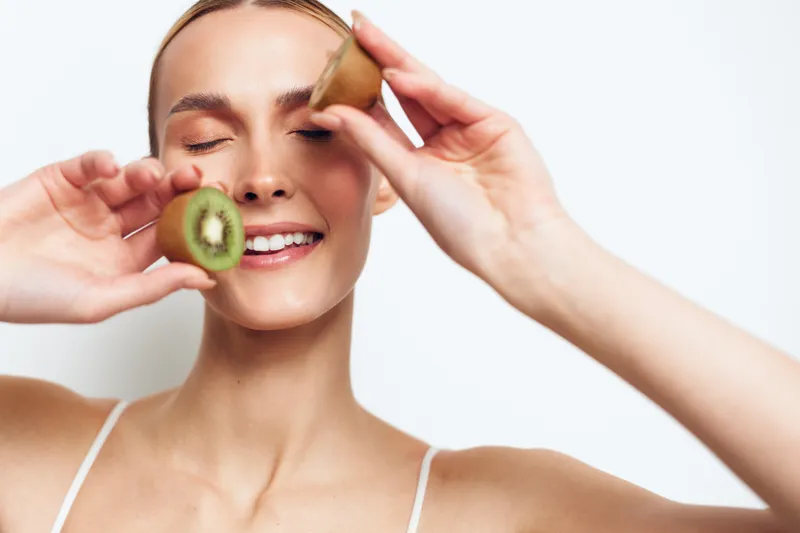 Happy woman holding kiwi slices near face, smiling with closed eyes, fresh fruit concept, healthy lifestyle, bright studio lighting, clean white background
