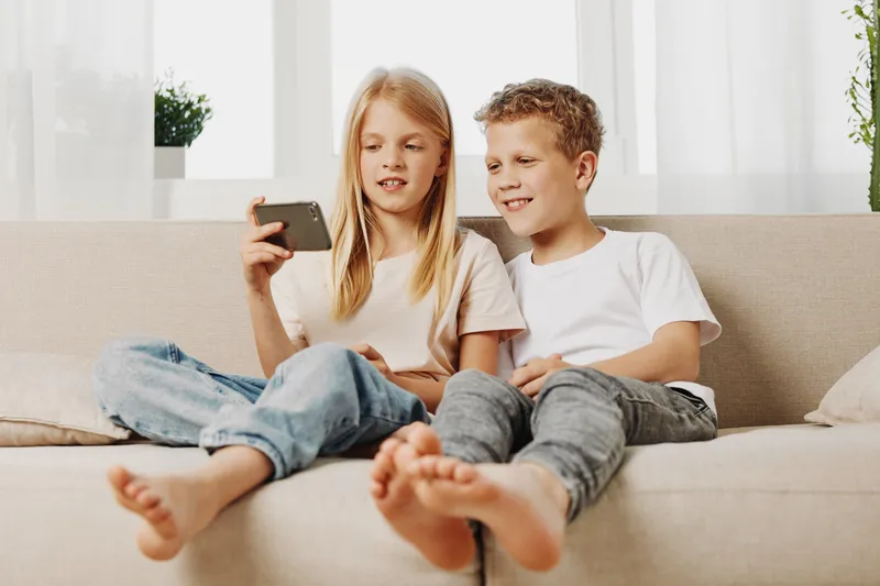 Two children sitting on a beige sofa looking at smartphone screen together enjoying casual moment in bright living room interior with natural light.