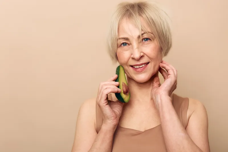 Smiling middle aged woman with short blonde hair holds avocado near face, showing natural beauty and healthy lifestyle. Concept of skincare, nutrition, wellness, and self care in studio.