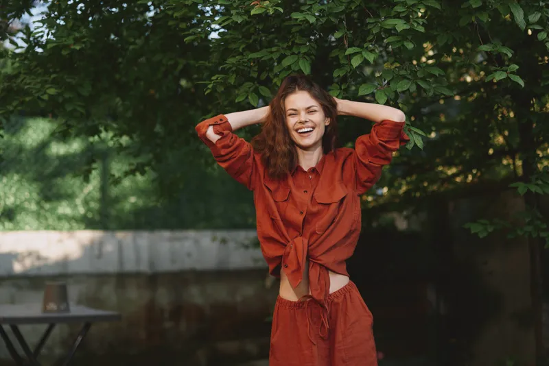 Cheerful woman outdoors, smiling with relaxed posture, lifestyle portrait in natural light, casual fashion outfit, sunlit greenery, carefree mood, vibrant colors