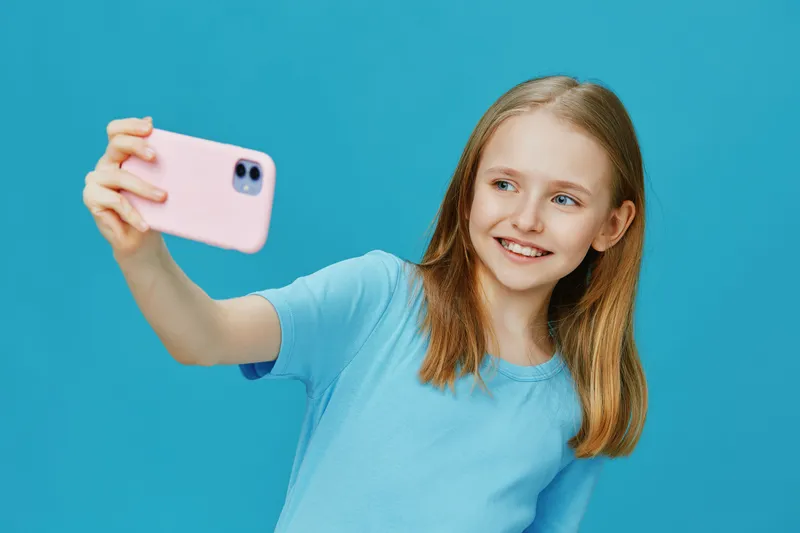 A cheerful girl taking a selfie with a smartphone against a vibrant blue background, showcasing happiness and youthfulness in casual attire