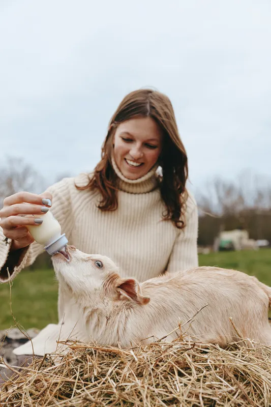 Cheerful woman outdoors feeding a goat with a bottle, cozy sweater, rustic farm setting, soft daylight, candid moment, gentle interaction, natural colors, countryside vibes, wholesome lifestyle