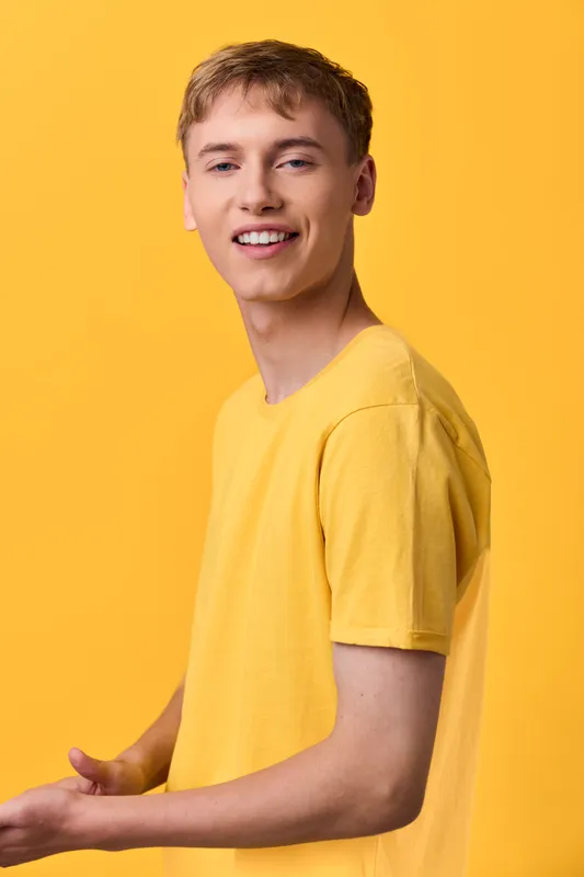 Smiling young man in a yellow shirt stands against a yellow background, presenting casual fashion, relaxed posture and friendly vibe for lifestyle or promotional content