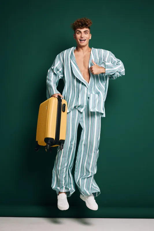 Young man in striped pajamas jumping with yellow suitcase on green background for travel, vacation and leisure. Casual outfit, happy expression and luggage for trip or holiday.