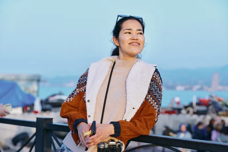 Outdoor, smiling female in a fur vest and casual jacket poses on a seaside boardwalk, enjoying sunny weather, coastal scenery, and a relaxed travel vibe.