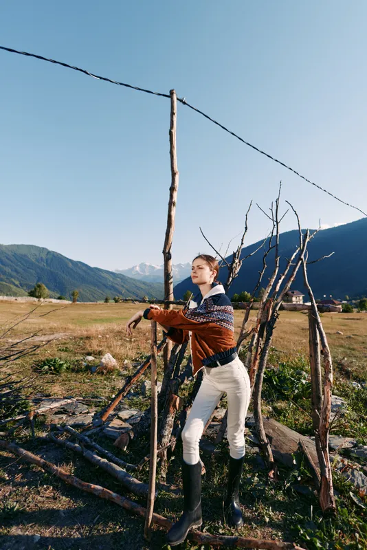 Woman countryside fence sticks mountains field: Young woman leans on rustic stick fence in open field with mountain valley backdrop, wearing sweater and boots for rural outdoor lifestyle.
