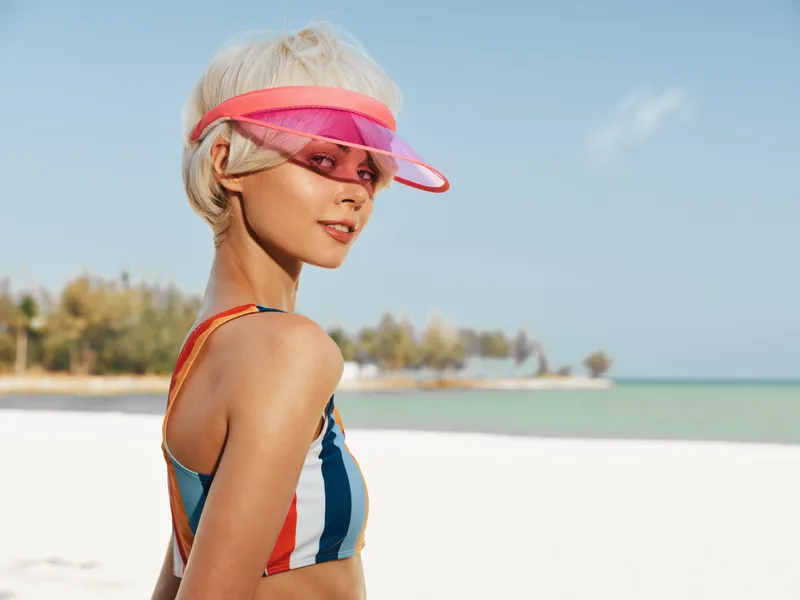 A stylish beach portrait features a smiling woman in a striped swimsuit, pink visor, and sunlit shoulders against clear blue sky and turquoise water.
