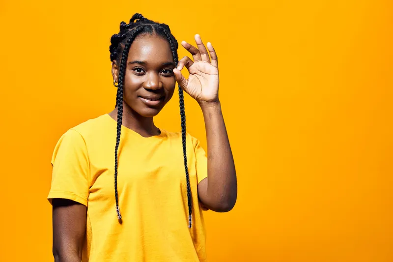 A young African woman in a bright yellow shirt smiling while making an OK gesture against a vibrant orange background, expressing confidence and positivity