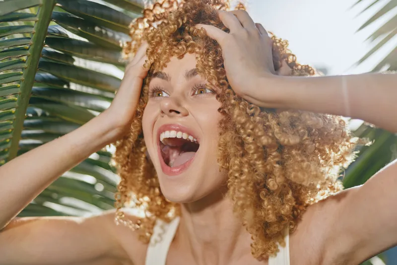 Excited woman with curly hair enjoying sunny day outdoors with tropical palm leaves background, expressing happiness and vibrant summer energy.
