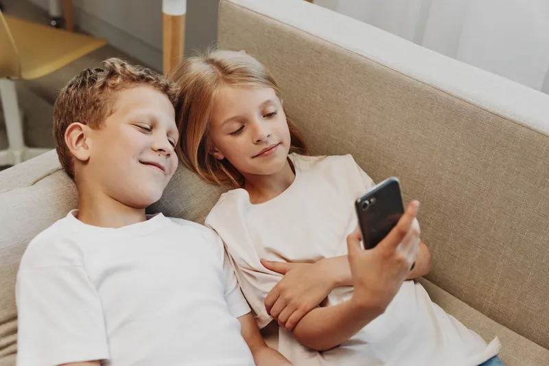 Two children lying on a sofa smiling and looking at a smartphone together. Casual home environment with natural light, concept of childhood, technology, and family bonding.