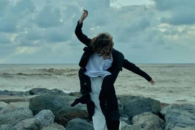 Movement, dance on rocky coastline with sea in background, wind swept coat and white clothing creating dynamic action, tension and a bold sense of motion in outdoor scenery