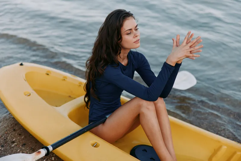A young woman sits in a yellow kayak by calm water, wearing a navy long sleeve top, looking thoughtful as she rests after paddling under soft sunlight.