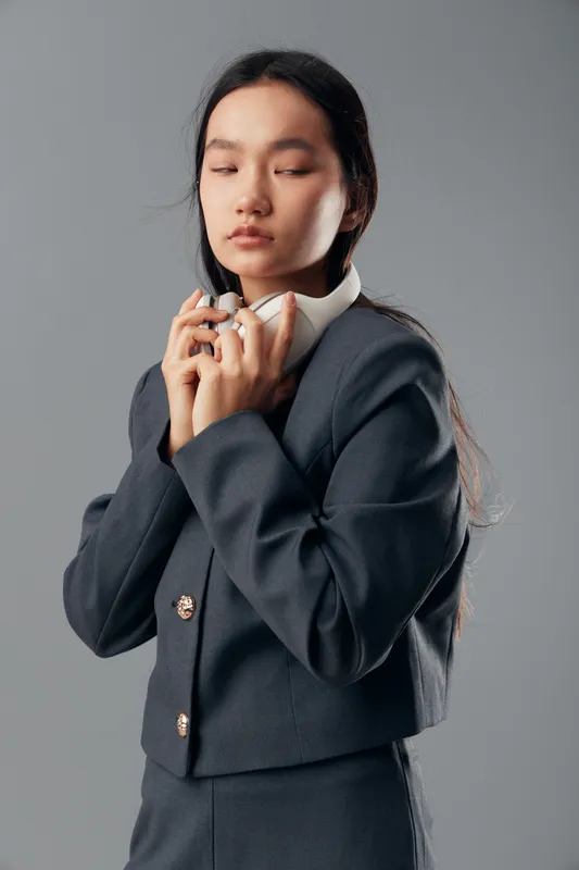 Young woman in dark formal suit holding white headphones with closed eyes and calm expression, standing against gray background, fashion portrait with serene mood and simple studio lighting.