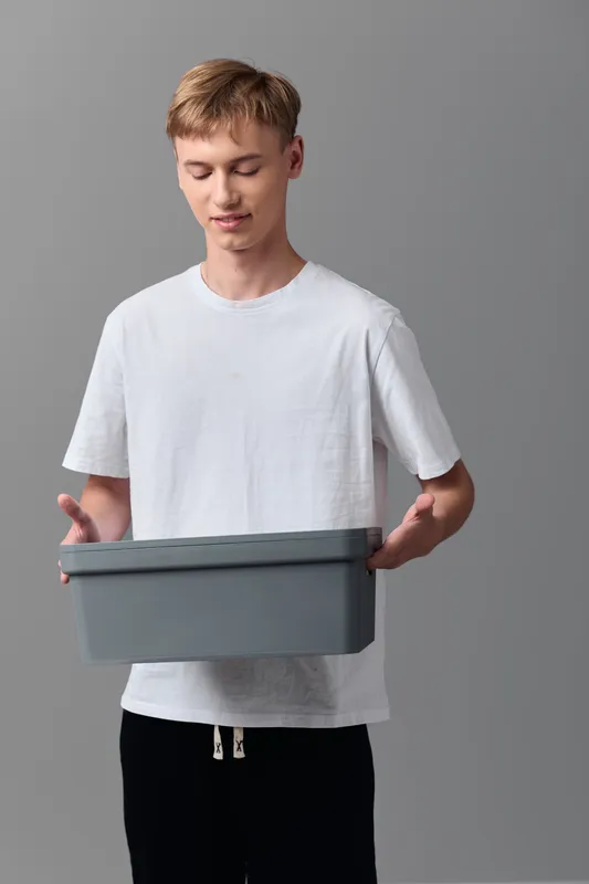 Casual young man holding a gray box against a neutral background, illustrating packaging and delivery concepts, simple logistics and stock handling in a clean studio setup