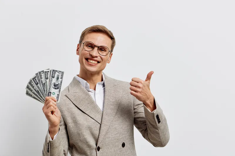 Smiling man in stylish beige suit and glasses holding dollar bills and giving thumbs up, celebrating financial success, studio portrait with plain white background, business and finance concept.