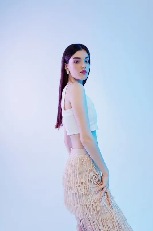 Young woman with long dark hair poses in a white top and fringed skirt against a soft blue background. Concept of beauty, fashion, and elegance in studio lighting.