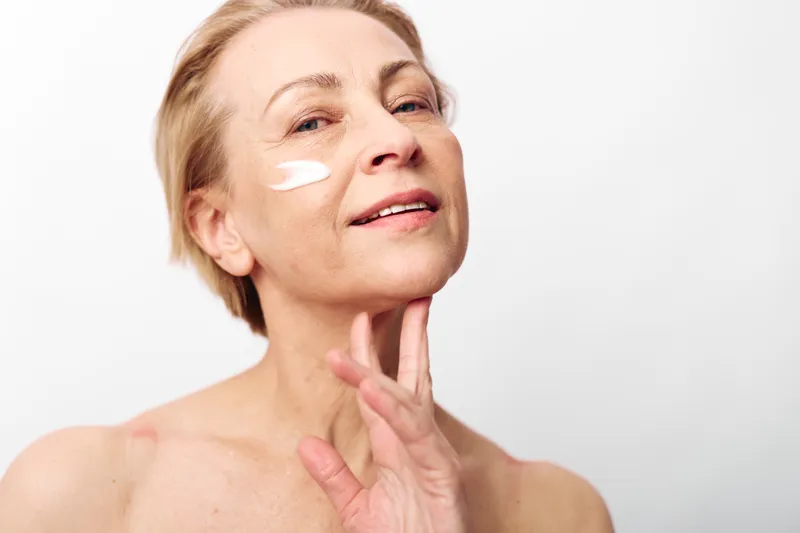 Mature woman applies facial cream on cheek with fingers, smiling gently on white background. Concept of skincare, self-care, beauty routine, and natural aging with healthy skin care.