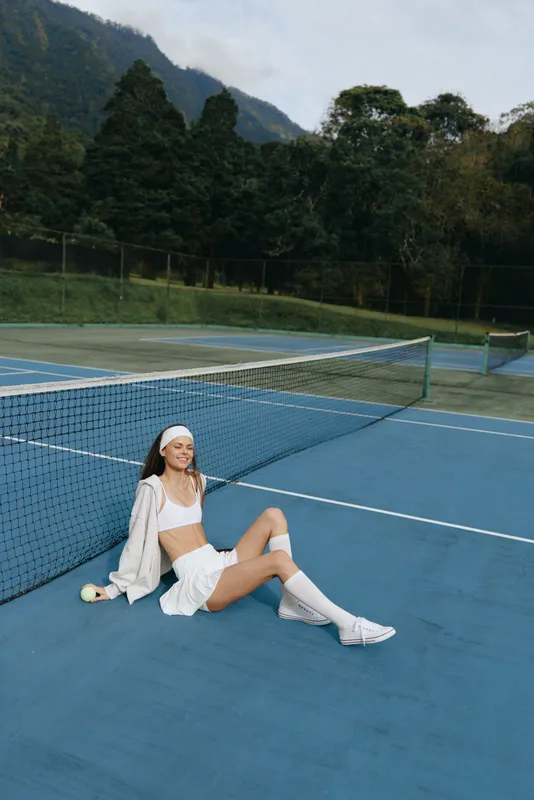 Young female tennis player in sporty attire exuding joy on a blue tennis court, wearing a headband, smiling and enjoying a sunny day outdoors