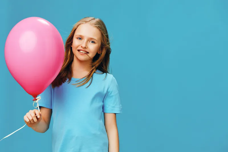 Happy girl with a pink balloon, wearing a blue t shirt against a bright blue background, evokes joy and innocence in a playful moment