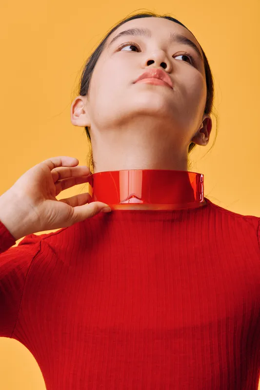 Young woman in red sweater adjusting red transparent choker necklace against yellow background. Minimalistic fashion portrait with modern accessories and clean style.