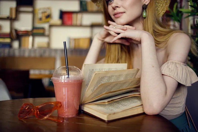 Женщина в шляпе сидит в кафе, перед ней стакан с напитком и книга для отдыха