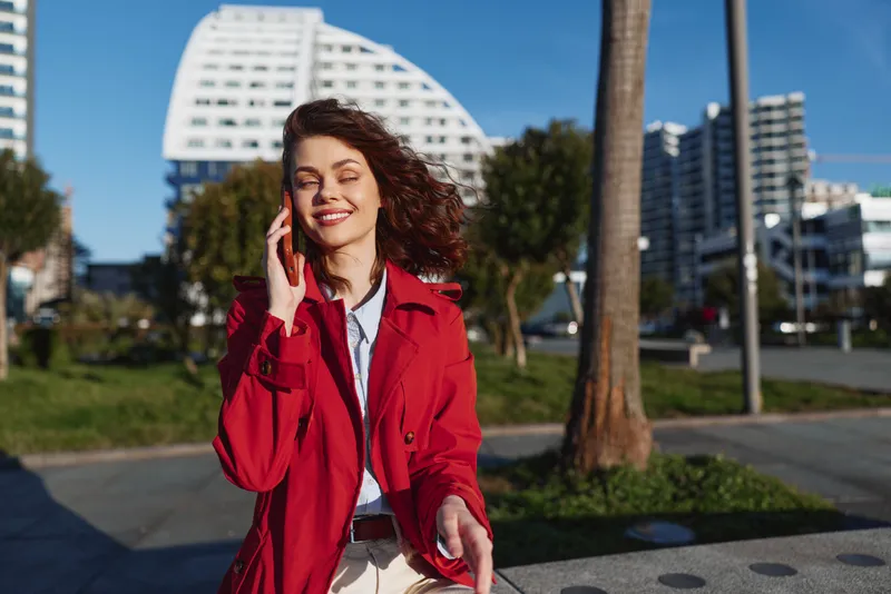 Smiling woman in a red coat talks on a phone outdoors, urban city street backdrop. Casual chic style with bright daylight and a confident, friendly mood.