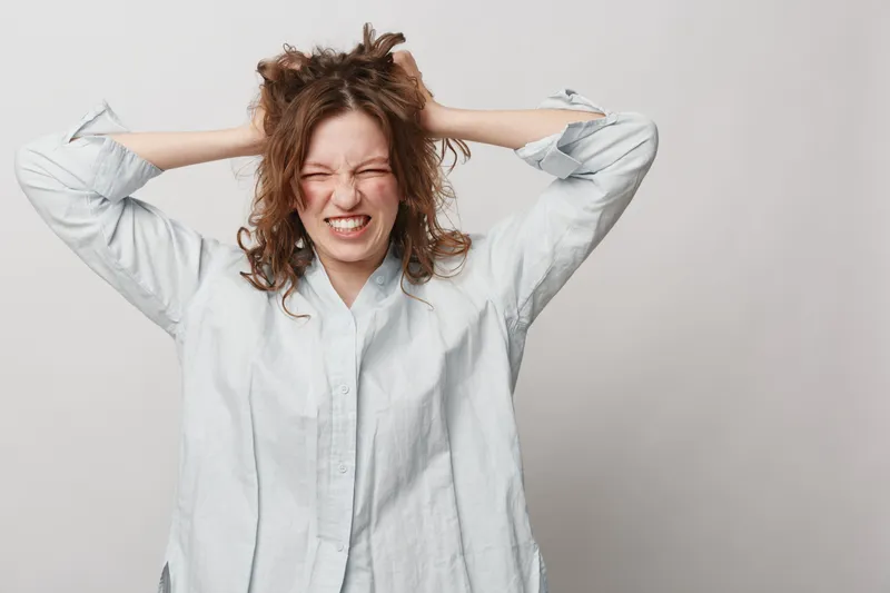 Frustrated young woman in white shirt pulls her hair and grimaces with closed eyes. Isolated on grey background. Concept of stress, anxiety, frustration, and emotional tension.