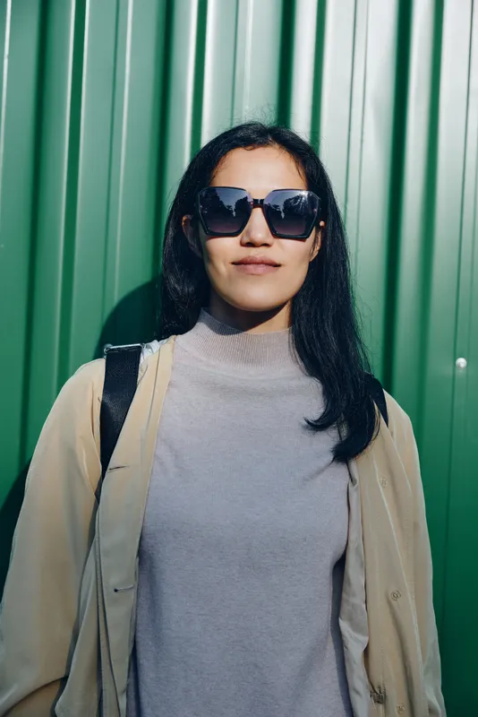 Woman wearing sunglasses against a green metal wall, casual sweater and jacket, urban street style, confident pose, sunny day, modern fashion mood