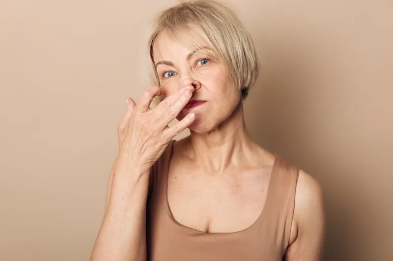Middle aged woman with short blonde hair wearing sleeveless top holds nose with fingers showing disgust on beige background. Concept of unpleasant smell and negative reaction.
