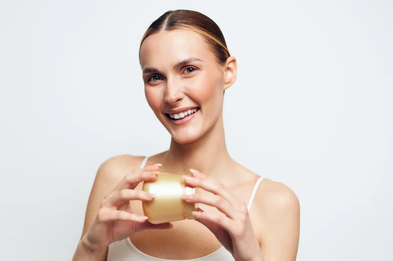 Smiling woman holding skincare cream jar, radiant complexion, natural beauty, clean white background, casual hairstyle, soft lighting, relaxed expression