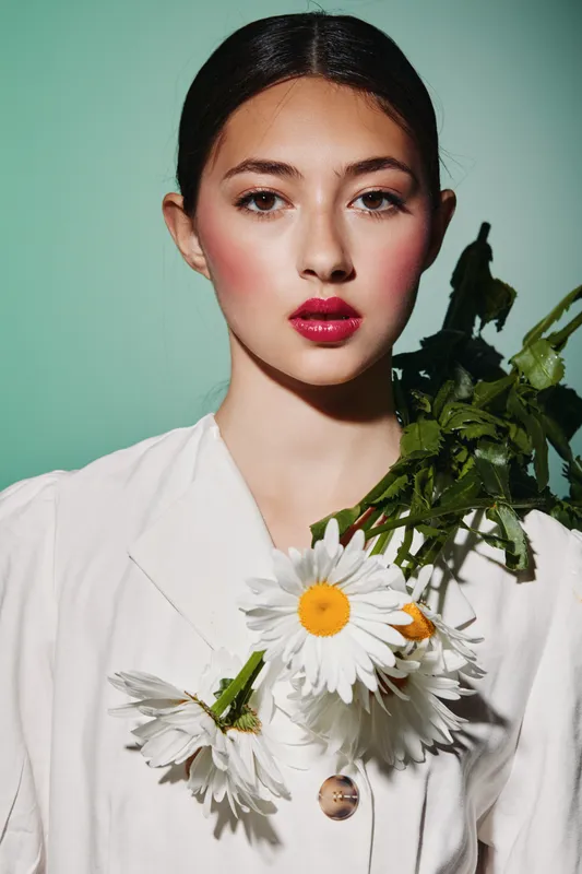 Young woman with glossy dark hair wears white jacket and holds large daisies close. Her bold red lips and shaved eyebrows create a high fashion beauty creative concept.
